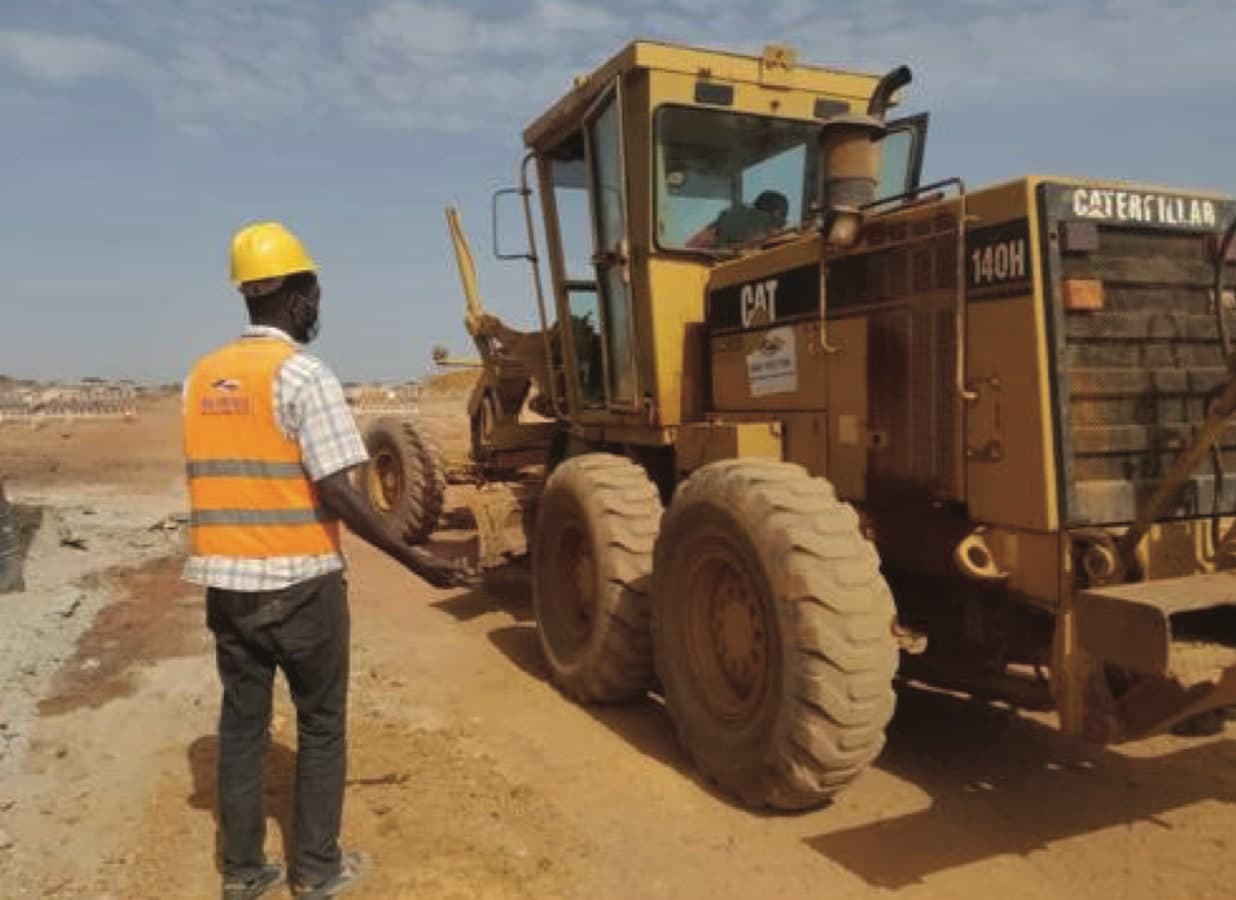 Chef de chantier en casque jaune guidant une niveleuse CAT 140H sur une voirie en terre.
